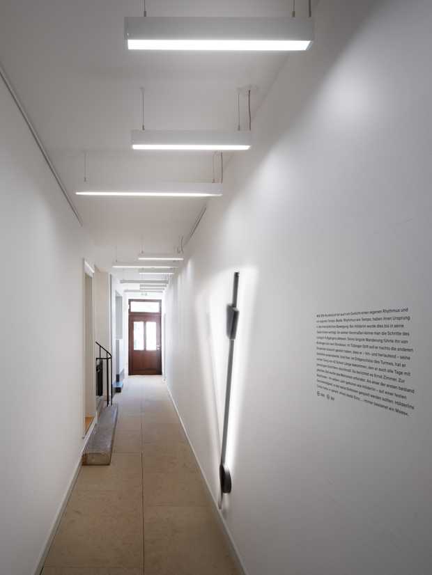Ein langer Gang mit weißen Wänden und einem Natursein-Boden. An der Decke hängen Lichtschienen. Ein großes schwarzes Pendel schwingt an der Wand hin und her.