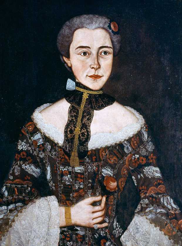 Hölderlins Mutter hat ein schmales Gesicht. Ihre Haare sind nach oben gesteckt. Sie trägt ein kostbares Kleid. Es hat ein Muster aus kleinen braunen und orangenen Blumen. Das Kleid ist oben ausgeschnitten und hat weiße Stickränder.