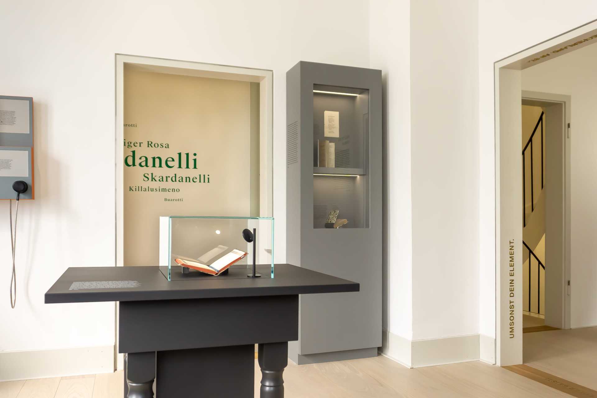 Raumansicht mit dunkler Tischvitrine in der Mitte und einer Vitrine mit Büchern an der rechten Wand. Durch die Türschwelle erkennt man einige der Pseudonyme, mit denen Hölderlin seine Turmgedichte unterzeichnete