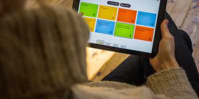 Auf einem Tablet sind bunte Kacheln in den Farben blau, grün, orange und gelb zu sehen. Eine Person wählt daraus die grüne Kachel mit der Aufschrift ›Der Frühling‹ aus.