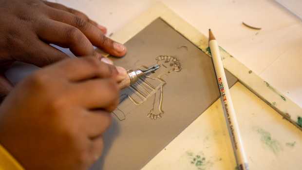 Jemand schnitzt mit der rechten Hand eine Figur in eine Linolplatte. Mit der linken Hand hält er die Platte fest.