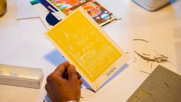 Eine Hand hält eine Postkarte in der Hand. Auf der Karte ist ein gelber Druck vom Hölderlinturm. Darunter steht das Wort 'sehen'.