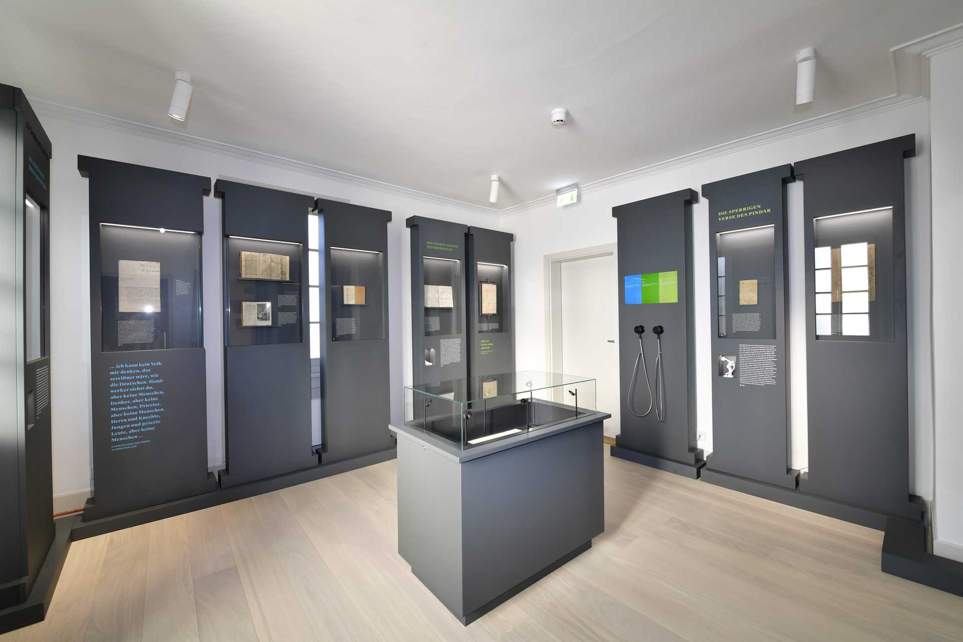 Raumansicht mit neun dunklen säulenartigen Vitrinen an der Wand und einer Vitrine mit Glashaube in der Mitte