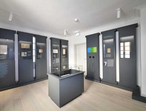 Raumansicht mit neun dunklen säulenartigen Vitrinen an der Wand und einer Vitrine mit Glashaube in der Mitte