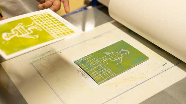 Eine grün eingefärbte Linolplatte mit einer Figur. Die Platte liegt auf einer Postkarte mit dem Wort 'Mensch'.