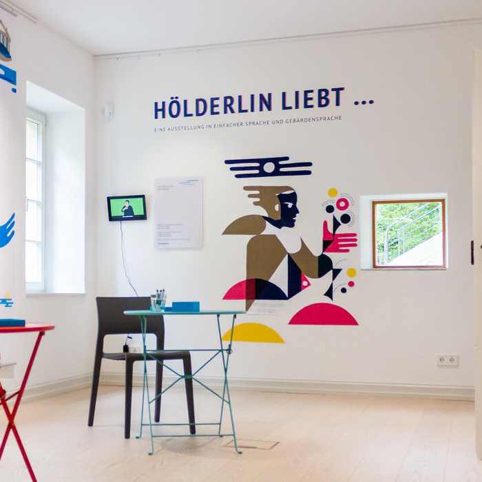 Ein Blick in die Sonderausstellung. Im Raum stehen zwei runde Tische. An den Wänden sind bunte Illustrationen und der Ausstellungstitel ›Hölderlin liebt...‹ angebracht.