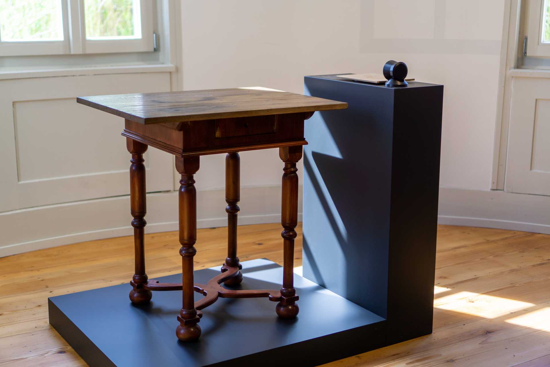 Schreibtisch mit Nussbaumfurnierter Zarge, gedrechselten Beinen und gekreuzten Stegen. Auf der Tischplatte aus Eiche befinden sich einige Tintenflecke und Abdrücke.