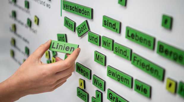 Eine Tafel mit Magnet-Wörtern aus Hölderlins Gedicht ›Die Linien des Lebens‹. Eine Hand hält das Wort ›Linien‹ zwischen Daumen und Zeigefinger.