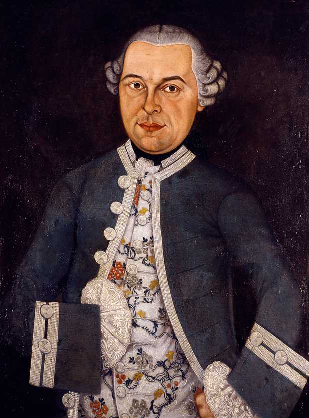 Hölderlins Vater hat ein rundes Gesicht. Er trägt eine graue Perücke mit hochgewickelten Locken. Er ist gekleidet mit einem blauen Jackett mit weißen Knöpfen. Darunter trägt er ein weißes Hemd mit aufgestickten Verzierungen in den Farben Gelb, Rot und Blau.